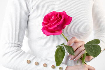 a girl in a white sweater with golden buttons on a white background holds a pink flower with a green stem in her hands with young white skin
