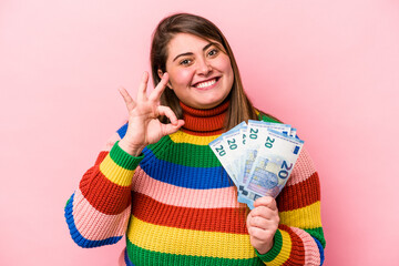 Young caucasian overweight woman holding banknotes isolated on pink background cheerful and confident showing ok gesture.