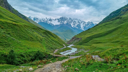Fototapeta na wymiar Patara Enguri River flowing down the a valley in the Greater Caucasus Mountain Range in Georgia, Svaneti Region, Ushguli. The Shkhara Mountain is covered by clouds. Snow-capped mountains.Bridge