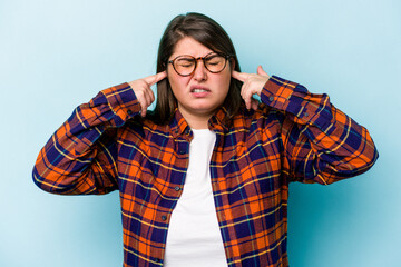 Young caucasian overweight woman isolated on blue background covering ears with fingers, stressed and desperate by a loudly ambient.