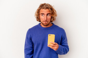Young caucasian man holding mobile phone isolated on white background confused, feels doubtful and unsure. - obrazy, fototapety, plakaty