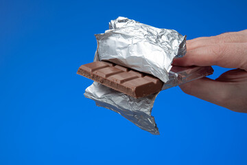 Generic Chocolate bar in aluminum foil held by male hand. Close up studio shot, isolated on blue...