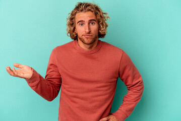 Young caucasian man isolated on blue background doubting and shrugging shoulders in questioning...