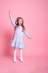 little girl in blue dress listening to music, dancing in big white headphones on pink background