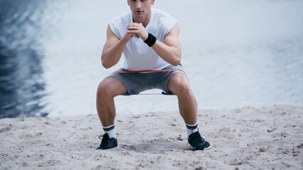 cropped view of athletic sportsman doing squat exercise on river shore