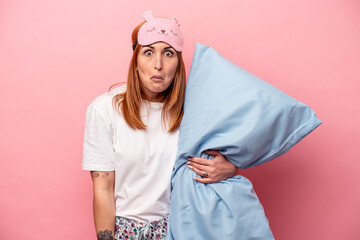 Young caucasian woman wearing a pajama holding pillow isolated on pink background shrugs shoulders...