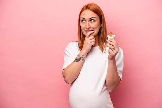 Young caucasian pregnant woman holding pacifier isolated on pink background relaxed thinking about something looking at a copy space.