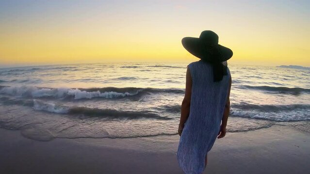 Attractive woman wearing dress and hat walking barefoot on the tropical beach at sunset