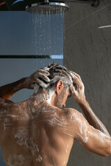 Back view of a man taking shower, washing his hair and body, standing under falling water drops in modern bathroom at home. Selfcare and wellness