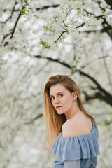 Naklejka premium Young pretty caucasian blonde woman with freckles wearing natural makeup in light blue dress near the beautiful blooming spring tree by the lake. Youth, freshness, beauty, happiness, emotions concept.