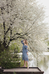 Fototapeta na wymiar Young pretty caucasian blonde woman with freckles wearing natural makeup in light blue dress near the beautiful blooming spring tree by the lake. Youth, freshness, beauty, happiness, emotions concept.