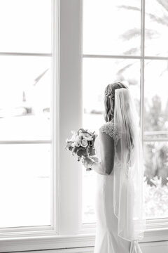 Bride with a bouquet of flowers stands near the window in the room Back view Black and white photo Bride morning preparation Bride in white wedding dress and veil standing near the window 