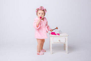 A cute little baby girl in a pink terry cloth and a curler robe, a mini dressing table and cosmetics is isolated on a white background, repeats the behavior of her mother