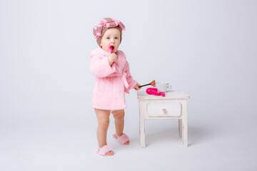 A cute little baby girl in a pink terry cloth and a curler robe, a mini dressing table and cosmetics is isolated on a white background, repeats the behavior of her mother