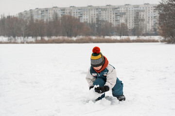 Boy sculpts snowman. Child plays with snow in winter park against the background of high-rise buildings. Games outside in winter.