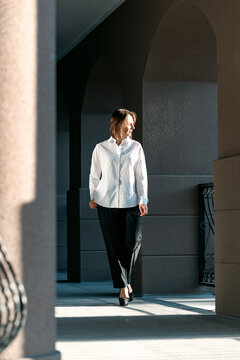 Young girl in white shirt and black pants poses outside building. Full-length portrait of woman in classic costume.