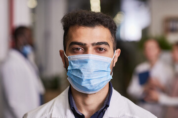 Young contemporary healthcare worker in mask and lab coat standing against his colleague having meeting in hospital corridor