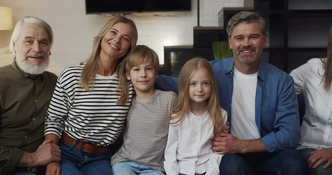 Portrait of Smiling Caucasian Big Family sitting on the sofa at Home. Cute Little Boy and Girl sitting Between Loving Parents and Grandparents. Hugging while looking into the camera. Three Generation