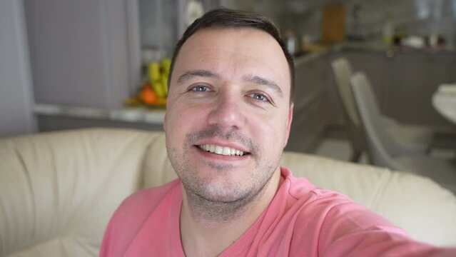 Image of millennial man smiling and taking selfie photo while sitting on sofa at home. Portrait of happy man in pink shirt taking selfie or having video call with friends showing new flat
