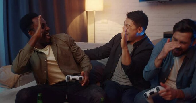 Happy Group of Diverse Men sitting playing Computer Games uses Gaming Joystick. Attractive Male Students spending time at Home with PlayStation. Have a Party. Cheering. Teamplay. Video Games.