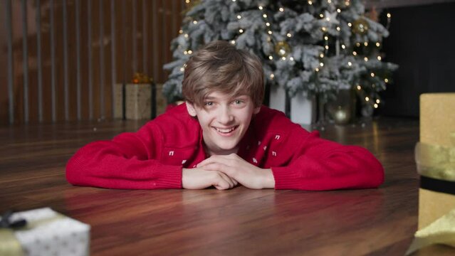 portrait of a joyful boy pushing beautiful gift boxes lying on floor background of Christmas tree, smiling and looking at camera