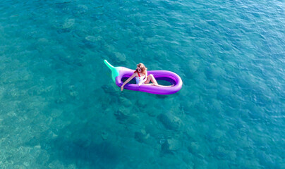 Young pretty woman in a white one piece swimsuit relaxing on a inflatable mattress in shape of big eggplant in turquoise sea water. Woman on air mtatress in transparent water.