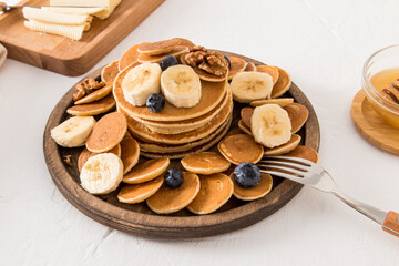 a large wooden plate with mini pancakes and a stack of thick pancakes in the middle. banana slices, fresh berries, nuts on them. white background.