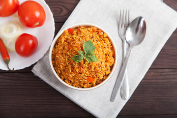 Jollof rice, tomatoes and hot peppers on a blue plate, fork, spoon on a linen napkin on a brown wooden background. National cuisine of Africa.