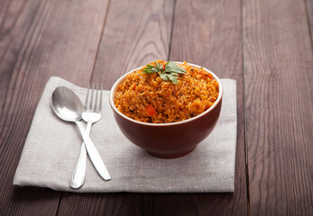 Jollof rice, tomatoes and hot peppers on a blue plate, fork, spoon on a linen napkin on a brown wooden background. National cuisine of Africa.