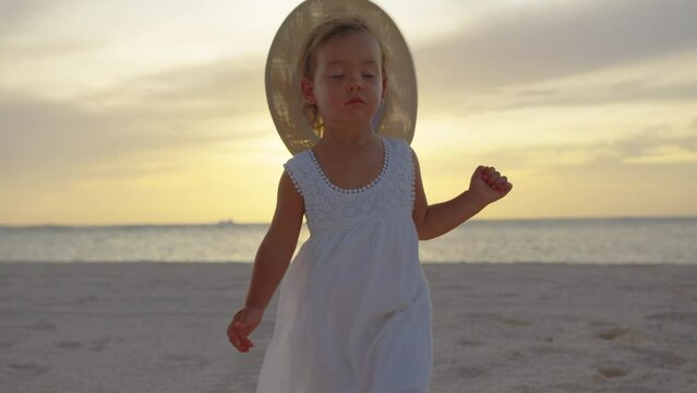 Happy little child girl in yellow hat walking at white sand during summer vacation on tropical beach near blue ocean. Smiling and cute toddler kid at the sea. Sunset rays in the background