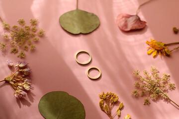 Gold wedding rings and various pressed flowers on pastel pink background, illuminated by sunlight....