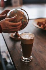 A glass glass of freshly brewed coffee with foam on the background of a globe. Men's hands hold a globe. Geography over a cup of latte