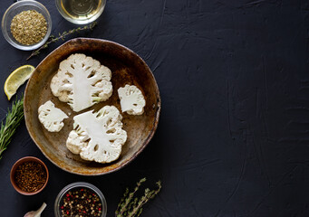 Cauliflower steak cooking. Raw cut cauliflower lie in a frying pan. Olive oil, herbs, various spices nearby. Dark background. Place for text.