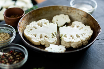 Cauliflower steak cooking. Raw cut cauliflower lie in a frying pan. Olive oil, herbs, various spices nearby. Dark background. Place for text.