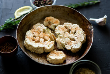 Cauliflower steak cooking. Raw cauliflower sprinkled with spices lies in a frying pan. Olive oil, herbs, various spices nearby. Dark background.