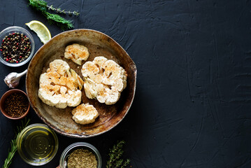Cauliflower steak cooking. Raw cauliflower sprinkled with spices lies in a frying pan. Olive oil, herbs, various spices nearby. Dark background. Copyspace.