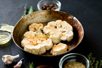 Cauliflower steak cooking. Raw cauliflower sprinkled with spices lies in a frying pan. Olive oil, herbs, various spices nearby. Dark background.