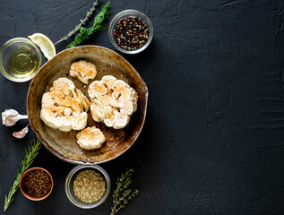 Cauliflower steak cooking. Raw cauliflower sprinkled with spices lies in a frying pan. Olive oil, herbs, various spices nearby. Dark background. Copyspace.
