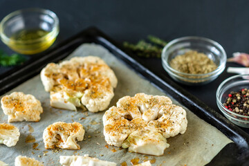 Cauliflower steak cooking. Raw cauliflower sprinkled with spices lies on a baking sheet. Olive oil, herbs, various spices nearby. Dark background.