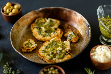 Cauliflower steak with spices lies in a frying pan. Olive oil, chimichurri sauce, capers, olives, herbs, various spices side by side. Dark background. Vegetarian food.