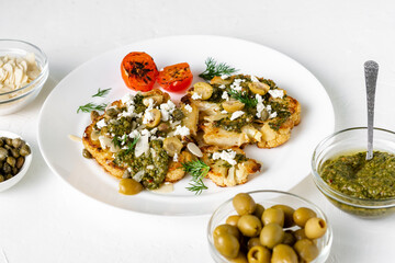 Cauliflower steak with spices, chimichurri sauce, almond flakes, olives, fried cherry tomatoes and capers on a white plate. Vegetarian food. White background.