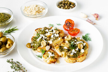 Cauliflower steak with spices, chimichurri sauce, almond flakes, olives, fried cherry tomatoes and capers on a white plate. Vegetarian food.