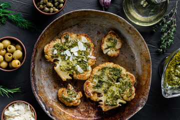 Cauliflower steak with spices lies in a frying pan. Olive oil, chimichurri sauce, capers, olives, herbs, various spices side by side. Dark background. Vegetarian food.