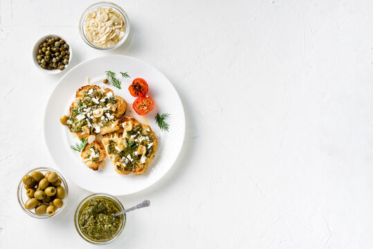 Cauliflower steak with spices, chimichurri sauce, almond flakes, olives, fried cherry tomatoes and capers on a white plate. Vegetarian food. White background. Place for text.