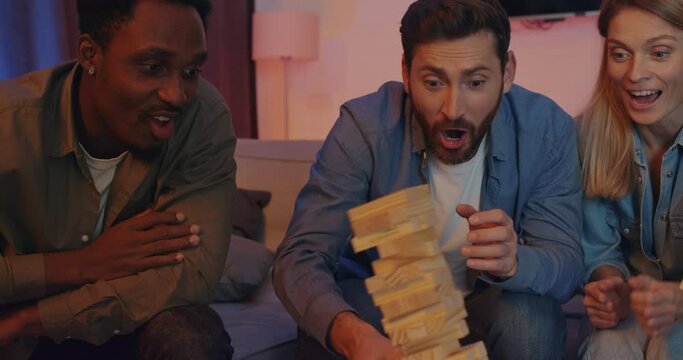 Cheerful Group of Friends of Men and Girls Playing Game Jenga at Home. Happy Friends Have Fun playing with Wooden Blocks in living room. While their friend accidentally Falls Down the Jenga Tower.