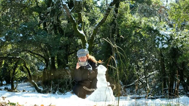 A woman is busy taking a picture of the snowman she has just created. a beautiful backlit, calm scene, at the snowy forest. a 4K video clip, January 21st, 2022, Golan Heights, Israel.