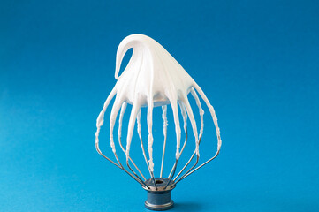 Whipped egg whites - beaten italian meringue on a wire whisk on blue background,  closeup and copy space.