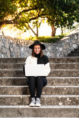 Una linda y alegre mujer caucásica con un sombrero 
sentada en las escaleras de un parque trabajando con su ordenador