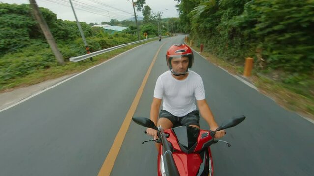 Male tourist motorcyclist in helmet and white T-shirt riding bike on intercity road in Phuket, Thailand. POV selfie, in front shot, on board camera, tropical forest all around 