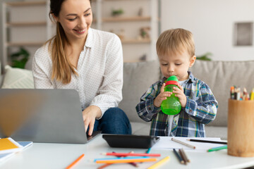 Happy millennial mom working on laptop while her adorable son drinking water from bottle, standing...